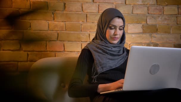 Nahaufnahme Porträt einer erwachsenen attraktiven Muslimin im Hijab mit dem Laptop, die einen Essay schreibt, während sie drinnen auf der Couch in einer gemütlichen Wohnung sitzt — Stockvideo