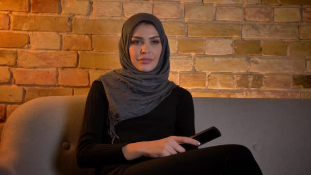 Κοντινό πλάνο του ενήλικα ελκυστικό μουσουλμανικό θηλυκό σε χιτζάμπ βλέποντας τηλεόραση ενώ κάθεται στον καναπέ σε εσωτερικούς χώρους σε ένα άνετο διαμέρισμα — Αρχείο Βίντεο