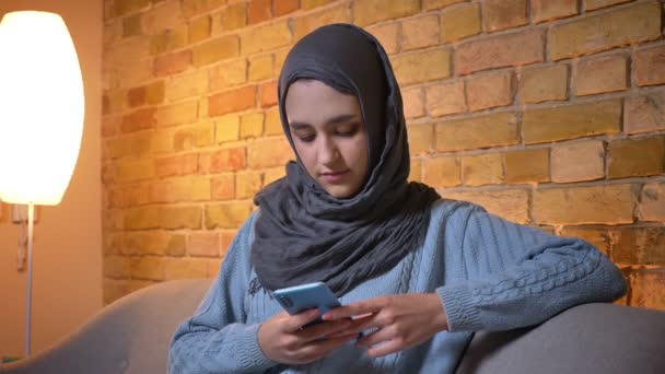 Sesión de primer plano de joven atractiva musulmana en hijab usando el teléfono y mirando a la cámara sonriendo mientras está sentado en el sofá en el interior de la acogedora casa — Vídeos de Stock