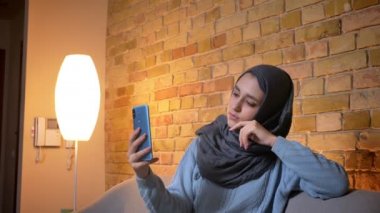 Rahat evde kanepede kapalı otururken başörtüsü telefonda selfie 'ler alarak genç çekici Müslüman kadın closeup ateş