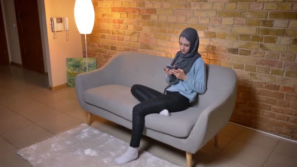 Gros plan pousse de jeune femme musulmane attrayante dans le hijab en utilisant le téléphone étant détendu sur le canapé à l'intérieur à la maison confortable — Video