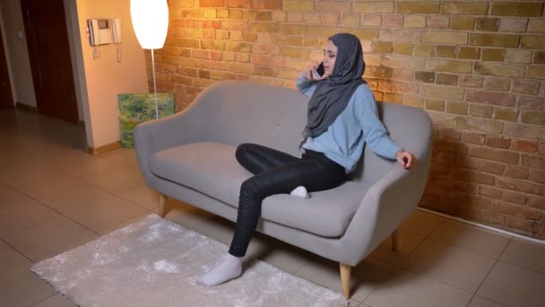 Κοντινό γύρισμα της νεαρής ελκυστική μουσουλμανική γυναίκα σε μαντίλα καλώντας στο τηλέφωνο, ενώ κάθονται στον καναπέ σε εσωτερικούς χώρους στο άνετο σπίτι — Αρχείο Βίντεο