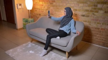 Sevimli genç çekici Müslüman kadın hijab telefonda selfie 'ler alarak ve kamera önünde poz rahat evde kanepede kapalı otururken çekim