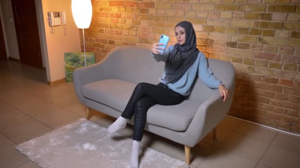 Nahaufnahme einer jungen attraktiven Muslimin im Hijab, die Selfies mit dem Handy macht und vor der Kamera posiert, während sie auf der Couch sitzt — Stockvideo