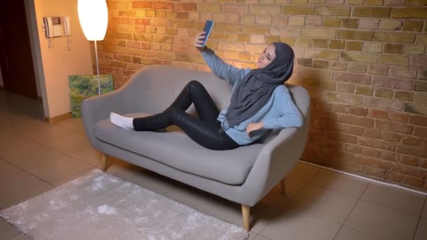 Крупним планом знімок молодої привабливої мусульманки в хіджабі робить селфі за допомогою телефону і позує перед камерою, сидячи на дивані — стокове відео