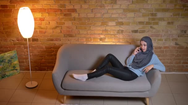 居心地の良い家に屋内のソファの上でのんびり休んで携帯電話を持つ若い魅力的なイスラム教徒の女性のクローズアップシュート — ストック動画