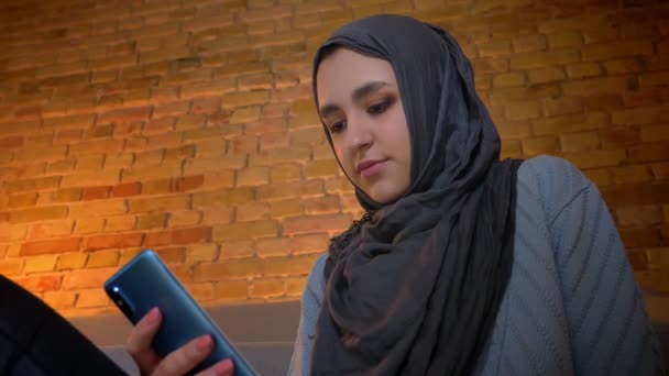 Närbild botten upp skjuta av unga attraktiva muslimska kvinnliga textning på telefonen och leende glatt medan du sitter på golvet inomhus på mysiga hem — Stockvideo