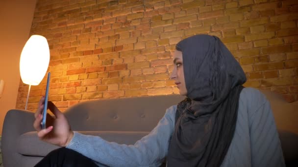 Närbild botten upp skjuta av unga attraktiva muslimska kvinnlig tonåring tar selfies på telefonen medan du sitter på golvet inomhus på mysiga hem — Stockvideo