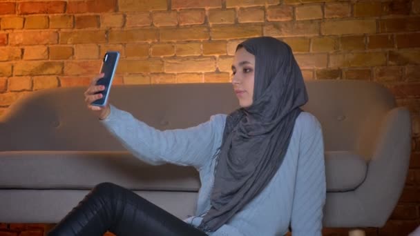 Gros plan du bas vers le haut pousse de jeune adolescente musulmane attrayante prenant selfies sur le téléphone et posant devant la caméra tout en étant assis sur le sol à l'intérieur à la maison confortable — Video