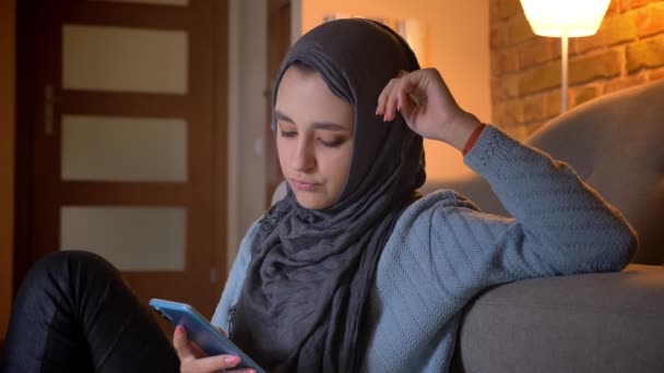 Tiro close-up de jovem fêmea muçulmana atraente em mensagens hijab no telefone enquanto sentado no chão na entrada da casa aconchegante — Vídeo de Stock