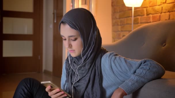 Gros plan shoot de jeune femme musulmane attrayante dans les vibrations en utilisant le téléphone en écoutant de la musique tout en étant assis sur le sol dans la porte à la maison confortable — Video