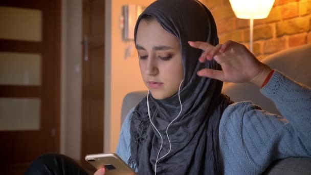 Rahat evde kapı katta otururken müzik ve sevinç ile şarkı dinlerken telefon kullanarak Vibes genç çekici Müslüman kadın closeup ateş — Stok video