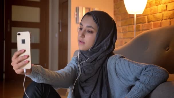 Κοντινό γύρισμα της νεαρής ελκυστική μουσουλμανική γυναίκα σε μαντίλα σε δονήσεις μιλούν μέσω βίντεο κλήση στο τηλέφωνο, ενώ κάθονται στο πάτωμα στην πόρτα στο άνετο σπίτι — Αρχείο Βίντεο