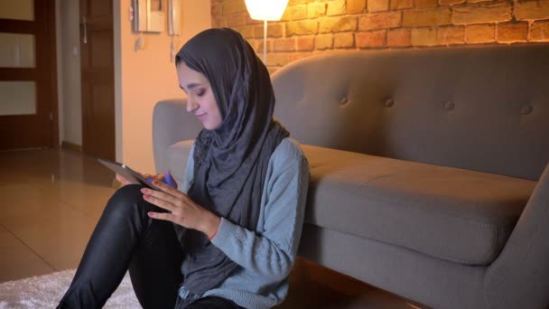 居心地の良い家で床の上に座っている間、若い魅力的なイスラム教徒の女性の笑顔と携帯電話でブラウジングのクローズアップ撮影 — ストック動画