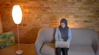 Dizüstü bilgisayar üzerinde hijab manifatura genç neşeli Müslüman kadın genç ve rahat evde kanepede kapalı otururken bir video çağrı sahip closeup ateş