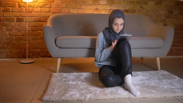 Tiro close-up de jovem fêmea muçulmana atraente em hijab sorrindo e rolando no telefone enquanto sentado no chão dentro de casa aconchegante — Vídeo de Stock