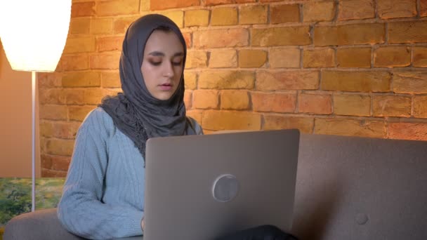 Primer plano de brote de joven atractiva musulmana hembra en hijab utilizando el ordenador portátil mientras está sentado en el sofá interior en casa acogedora — Vídeo de stock