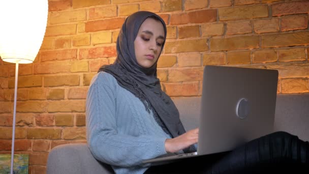 Primer plano de brote de joven atractiva estudiante musulmana en hijab utilizando el ordenador portátil y estudiar en línea mientras está sentado en el sofá en el interior de la acogedora casa — Vídeo de stock