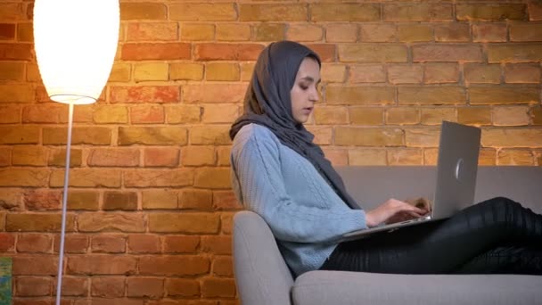 Close-up vista lateral atirar de jovem atraente muçulmano fêmea no hijab usando o laptop enquanto sentado no sofá dentro de casa aconchegante — Vídeo de Stock