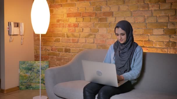 Closeup oldalmegtekintéshez lő fiatal vonzó muszlim nő hidzsáb segítségével a laptop és a munka otthonról ülve a kanapén beltérben egy hangulatos lakás — Stock videók