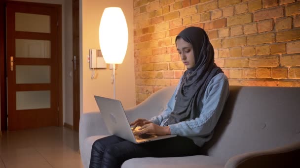 Κοντινό γύρισμα της νεαρής ελκυστική μουσουλμανική θηλυκό έφηβο σε μαντίλα χρησιμοποιώντας το φορητό υπολογιστή και έχοντας μια βιντεοκλήση, ενώ κάθονται στον καναπέ μέσα στο άνετο σπίτι — Αρχείο Βίντεο