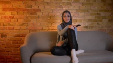 Rahat evde kanepede kapalı otururken TV izlerken ve uzaktan kumanda tutarak hijab genç çekici Müslüman kadın closeup ateş