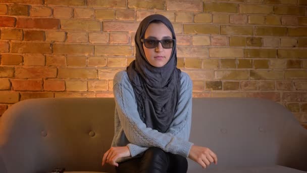 Sesión de primer plano de joven atractiva musulmana en hijab viendo una película en 3D en la televisión y manteniendo el control remoto mientras se sienta en el sofá en el interior de la acogedora casa — Vídeos de Stock