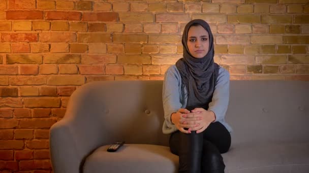 Κοντινό γύρισμα του νεαρού ελκυστικό μουσουλμανικό θηλυκό σε μαντίλα βλέποντας μια θλιβερή ταινία στην τηλεόραση, ενώ κάθονται στον καναπέ μέσα στο άνετο σπίτι — Αρχείο Βίντεο