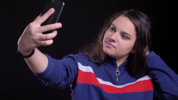 Primer plano retrato de adulto atractivo caucásico hembra tomando selfies en el teléfono y posando delante de la cámara con fondo aislado en negro — Vídeos de Stock