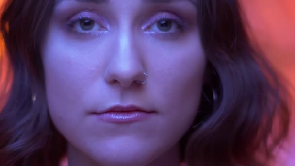 Zbliżenie strzelać młodych atrakcyjnych kaukaski kobiece twarz z piercing nosa i piękne oczy patrząc na aparat z neonowym czerwonym tle — Wideo stockowe