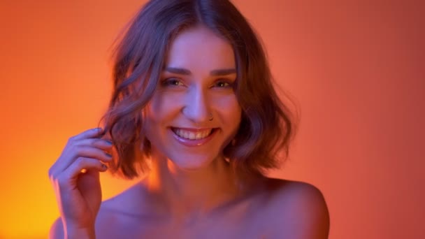 Primo piano sparare di giovane attraente femmina caucasica con spalle nude e giocare con i capelli guardando la fotocamera con sfondo rosso neon — Video Stock