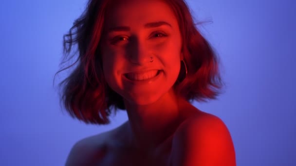 若い魅力的な白人の女性のクローズアップの撮影は幸せにネオン青と赤の背景を持つカメラを見て笑う — ストック動画