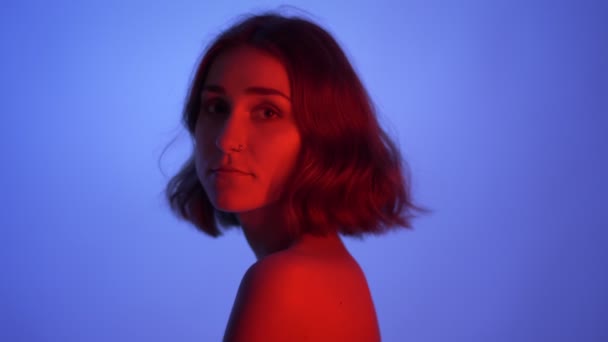 Closeup side view shoot of young sexy female with naked shoulders looking at camera and smiling with neon blue and red background — Stock Video