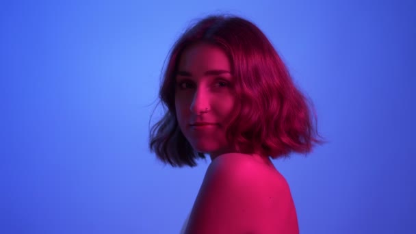Kamera bakıyor ve Neon mavi ve pembe arka plan ile baştan çıkarıcı gülümsüyor çıplak omuzlar ile genç seksi kadın closeup yan görünüm çekimi — Stok video