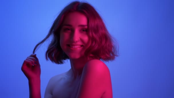 Closeup side view shoot of young sexy female with naked shoulders looking at camera and playing with her hair with neon blue and pink background — Stock Video