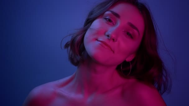 Съемки крупным планом молодой сексуальной женщины, счастливо улыбающейся, глядя в камеру с неоновым голубым фоном — стоковое видео