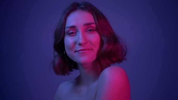 Closeup shoot of young pretty female smiling posing in front of the camera with her hair fluttering with neon blue background — Stock Video