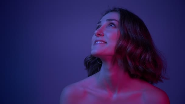 Primo piano sparare di giovane bella femmina con spalle nude sorridente posa davanti alla fotocamera con i capelli svolazzanti con sfondo blu neon — Video Stock