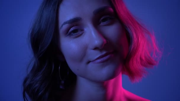 Close-up zijaanzicht schieten van jonge schattige vrouwelijke gezicht glimlachend gelukkig draaien om de camera met neon blauwe achtergrond — Stockvideo