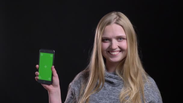 Porträt des blonden glatthaarigen Mädchens, das den grünen Bildschirm des Smartphones demonstriert, um die App auf schwarzem Hintergrund zu beraten. — Stockvideo