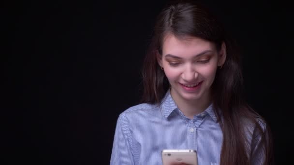 Porträt einer jungen brünetten Geschäftsfrau in blauer Bluse, die vor schwarzem Hintergrund lächelnd in ihr Smartphone schaut. — Stockvideo