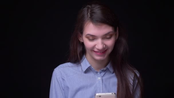 Urocza brunetka biznesmen w niebieskiej bluzce uśmiechając oglądanie w smartfonie na czarnym tle. — Wideo stockowe