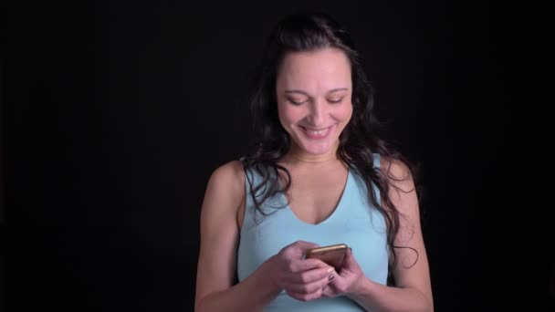 Retrato de mujer de mediana edad mirando alegremente en el teléfono inteligente y radiante en la cámara sobre fondo negro . — Vídeos de Stock