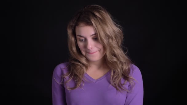 Portret van cute blonde Kaukasische vrouw glimlachend schuchter in de camera op zwarte achtergrond. — Stockvideo