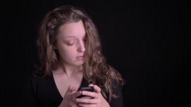 Akıllı telefona dikkatlice izlerken ve siyah arka planda sürpriz gösteren genç kıvırcık saçlı kızın portresi.