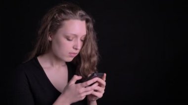 Akıllı telefona ciddiye izlerken genç kıvırcık saçlı kız portre ve siyah arka planda kameraya gülümsüyor.