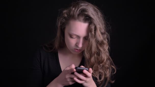 Porträtt av unga Curly-haired flicka tittar på allvar i smartphone på svart bakgrund. — Stockvideo