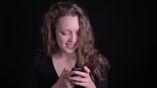 Retrato de una joven de pelo rizado mirando con alegría en un smartphone sobre fondo negro . — Vídeos de Stock