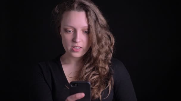 Portret van jonge krullend-haired meisje praten vreugdevol in videochat met behulp van smartphone op zwarte achtergrond. — Stockvideo
