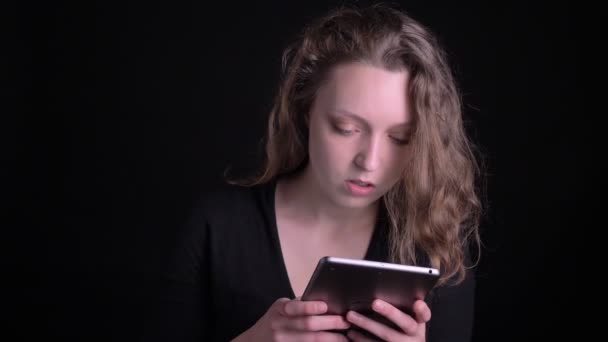 Portrait de jeune fille aux cheveux bouclés impliqué regardant sérieusement en tablette sur fond noir . — Video
